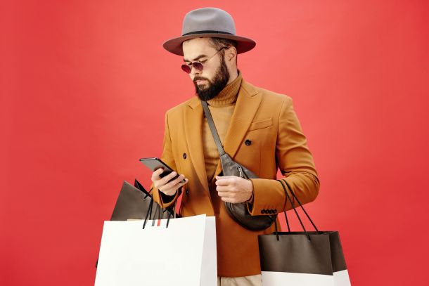 Foto von Max Fischer von Pexels, das eine Person mit Hut, Sonnenbrille, Pulli und Sakko zeigt, die Einkaufstüten trägt und auf ihr Smartphone schaut.