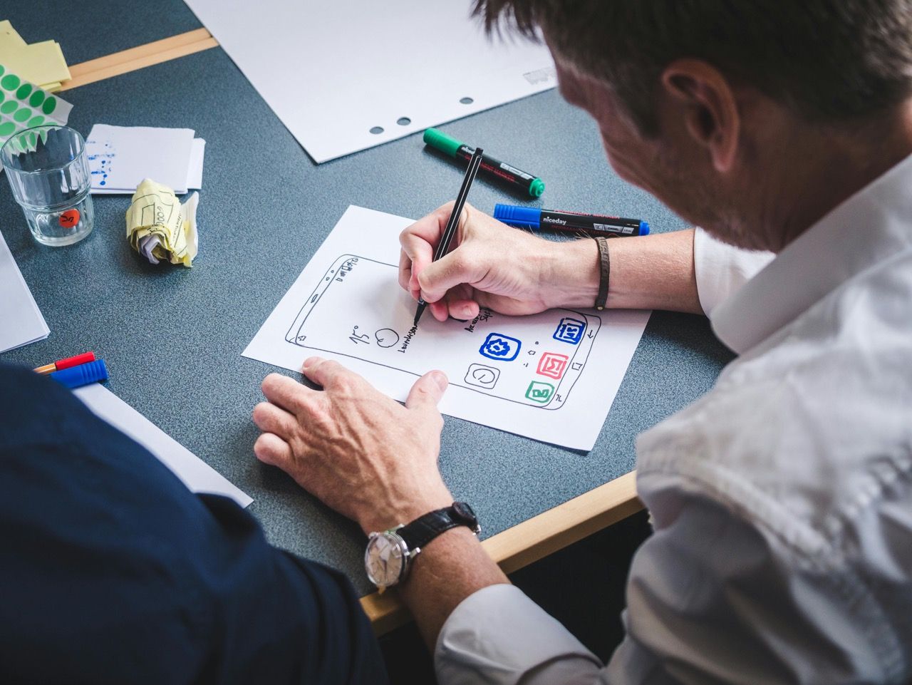 Two people are working at a table on a hand-drawn app design on paper, surrounded by pens and notes.