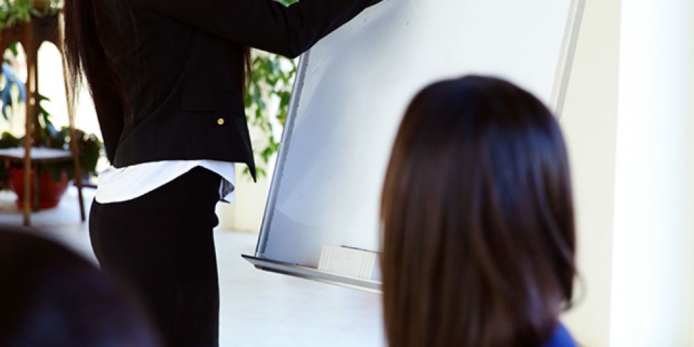 Eine junge Frau in einem Blazer präsentiert vor einer kleinen Gruppe an einem Flipchart.