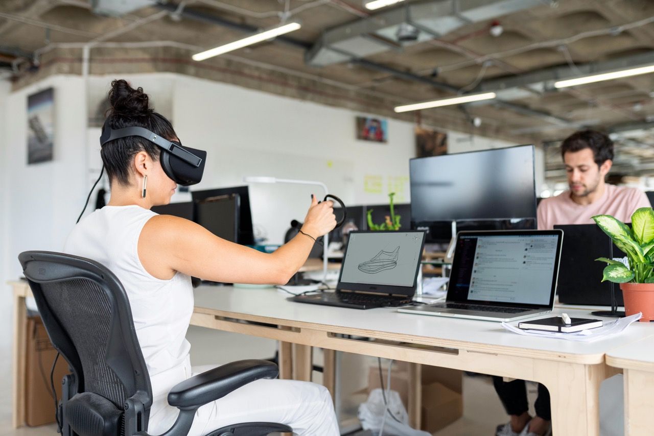 A product designer sits in an open-plan office, has two laptops in front of her and is currently using a VR headset with controllers.