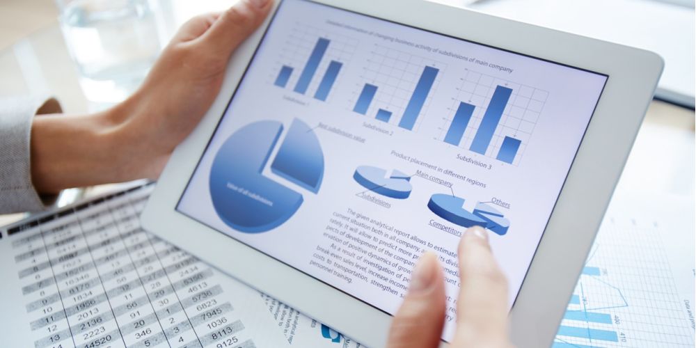 A person holding a tablet on which graphs are displayed above printouts with tables.