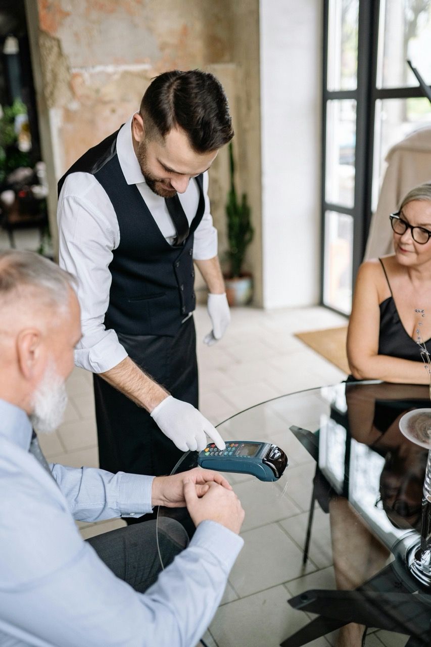 A waiter in an upscale restaurant helps an elderly couple pay with a card reader at the table.