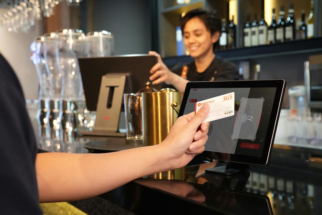 A customer holds a credit card against a terminal to make a contactless payment in a bar, with a smiling bartender in the background.