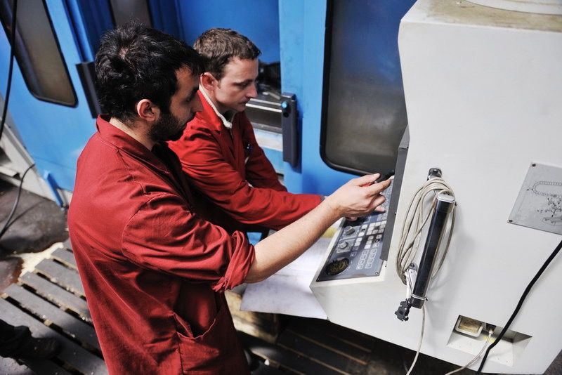 Two people are standing in front of the control panel of a production machine, discussing something.