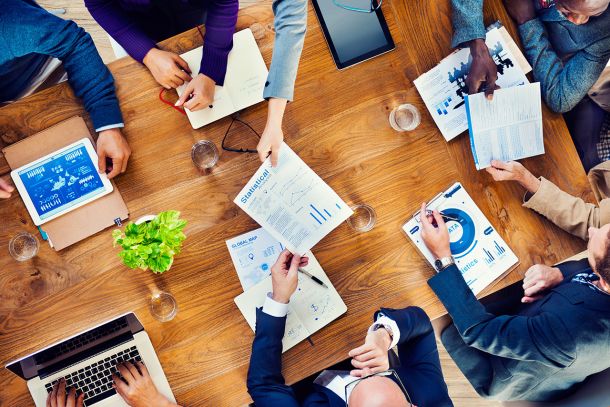 Personen sitzen mit Laptops, Tablets und Papier Dokumentan an einem Tisch.