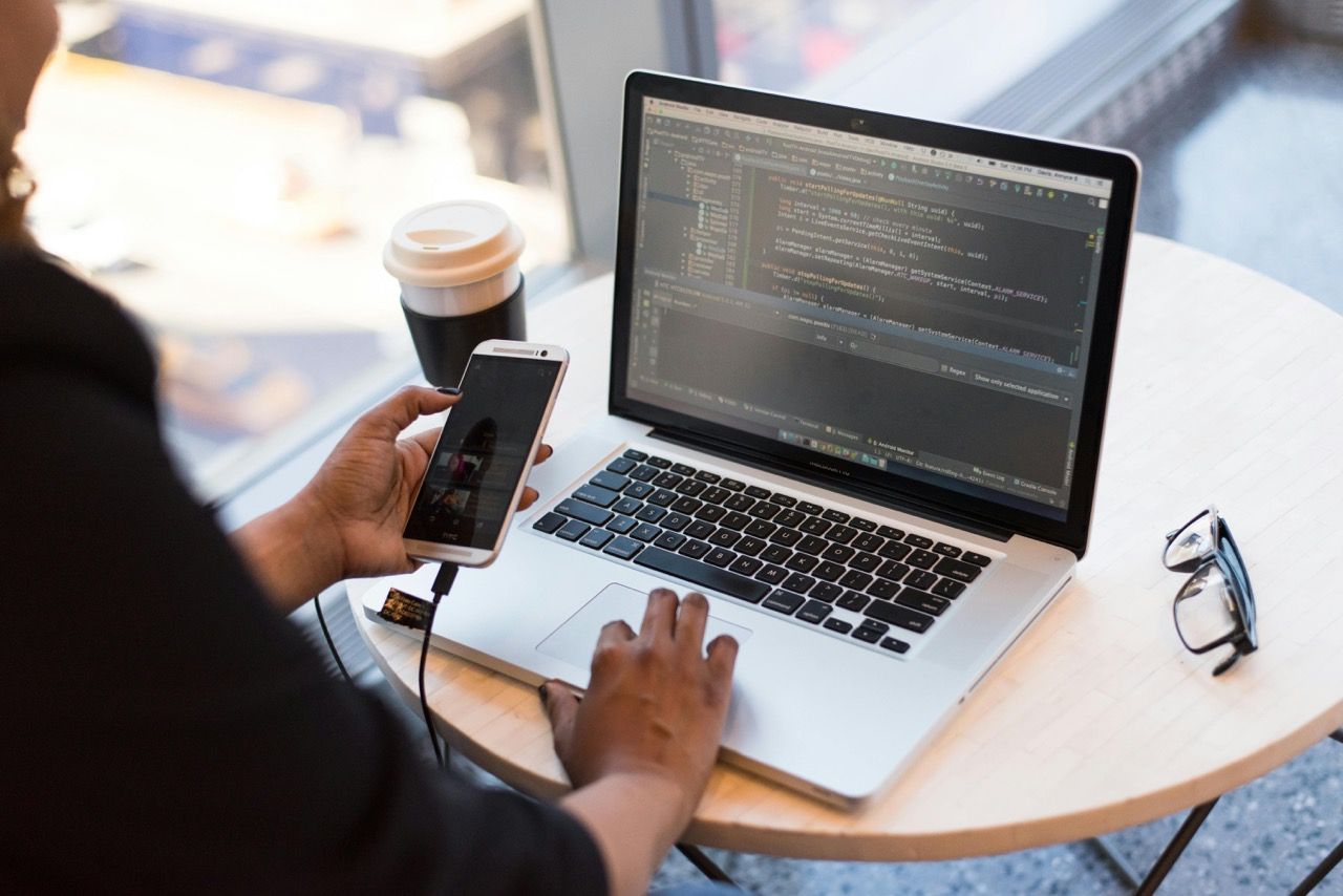 Developer sits with a coffee at the laptop and tests an app on the connected smartphone.