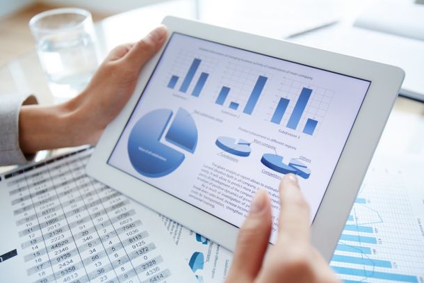 A person holding a tablet on which graphs are displayed above printouts with tables.+