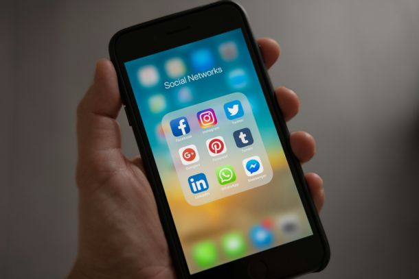 A hand holding an old iPhone, the display showing the home screen with the Social Networks folder open, containing app icons for Facebook, Instagram, and more.