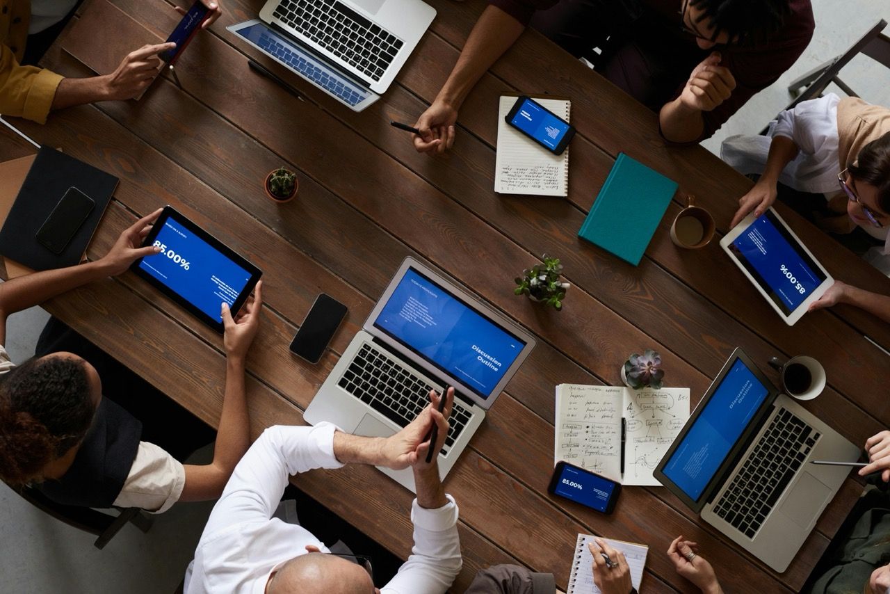 A top view of a group of people sitting at a conference table with laptops, tablets and smartphones.